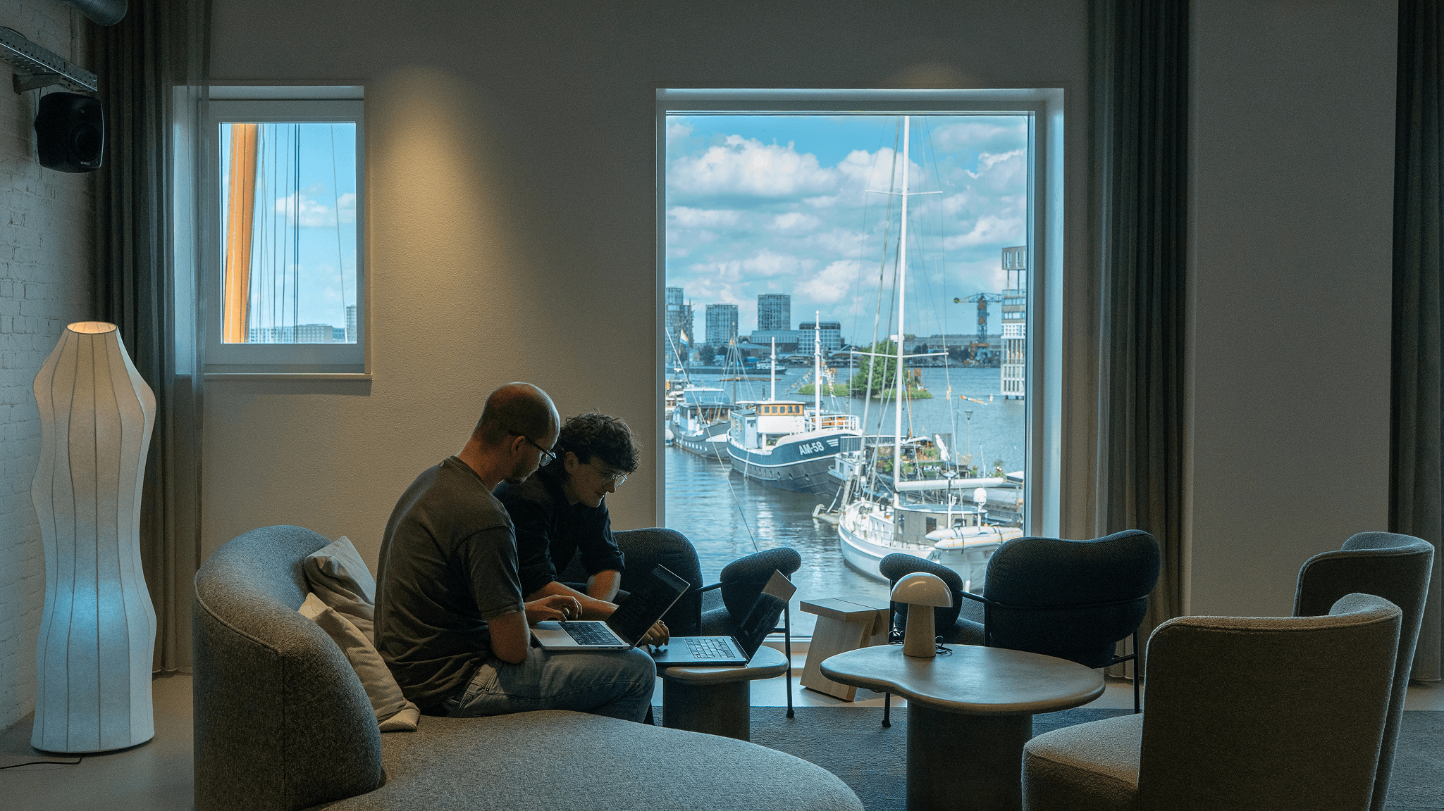 Two colleagues collaborating on laptops in Eidra's Amsterdam office, with a view of the harbour and sailboats through large windows.