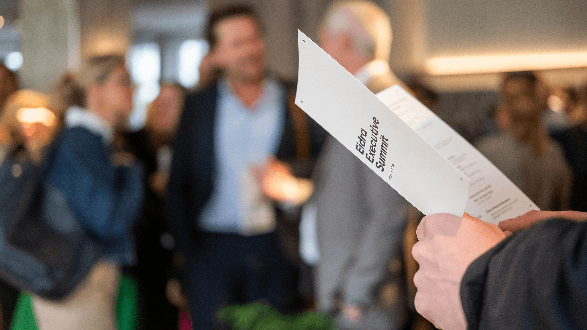 A hand holds an Eidra Executive Summit programme while attendees network in the background.
