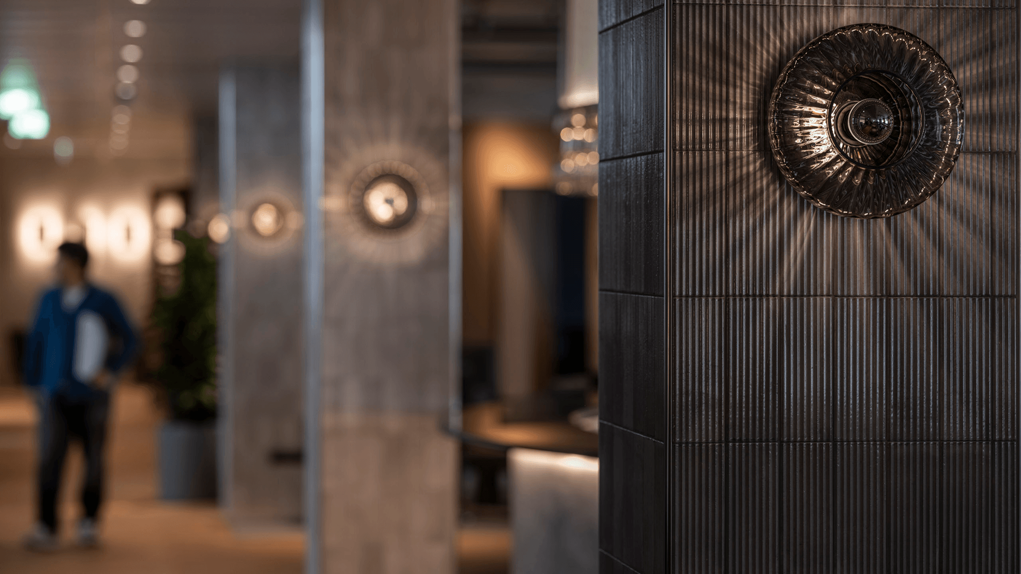 A close-up of an ornate wall light on dark ribbed tiles in an Eidra office, with a softly blurred corridor and a person walking in the background.