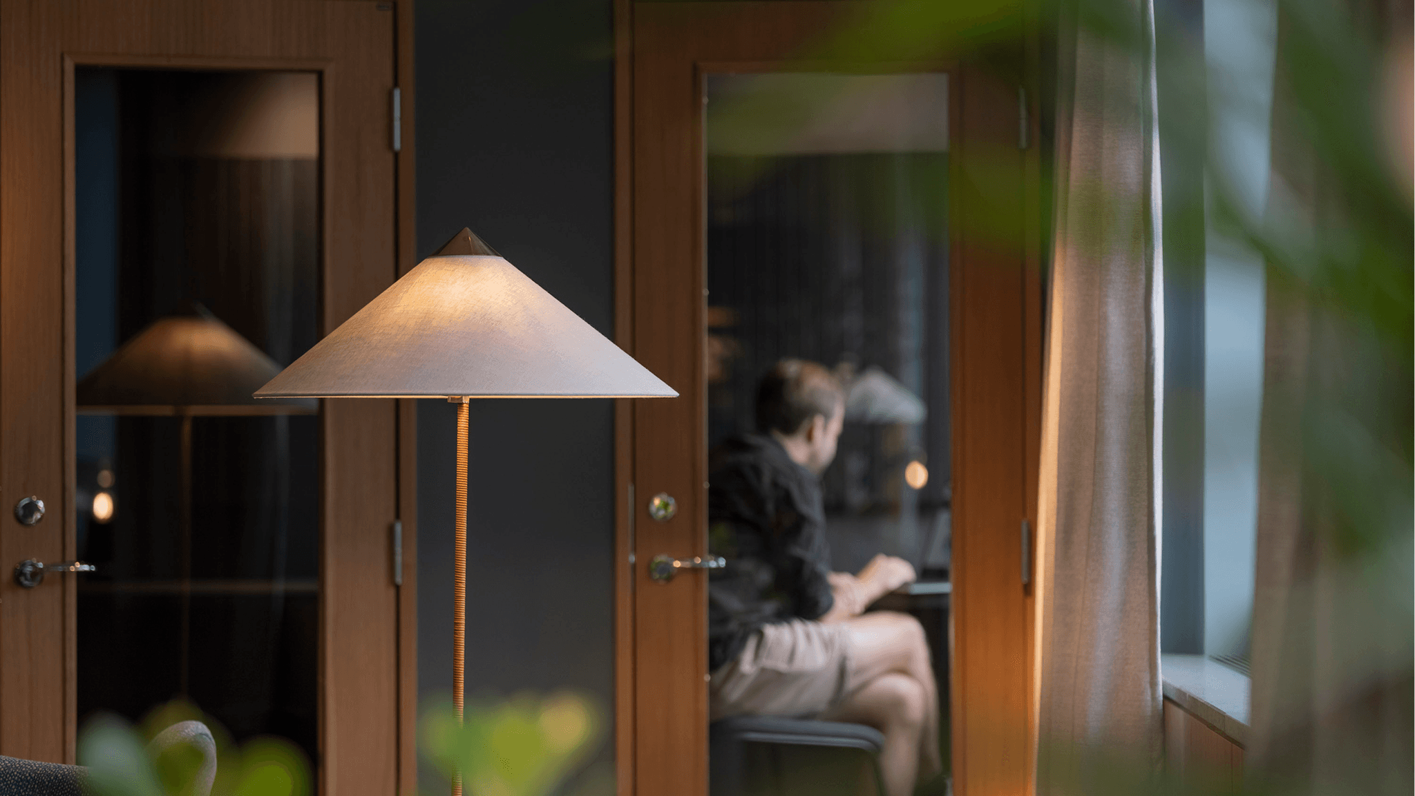 A designer table lamp in sharp focus in front of a warmly lit meeting pod, with a person working inside in the background.